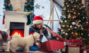 Woman wearing a santa hat opening a gift next to the Christmas tree with her small dog.